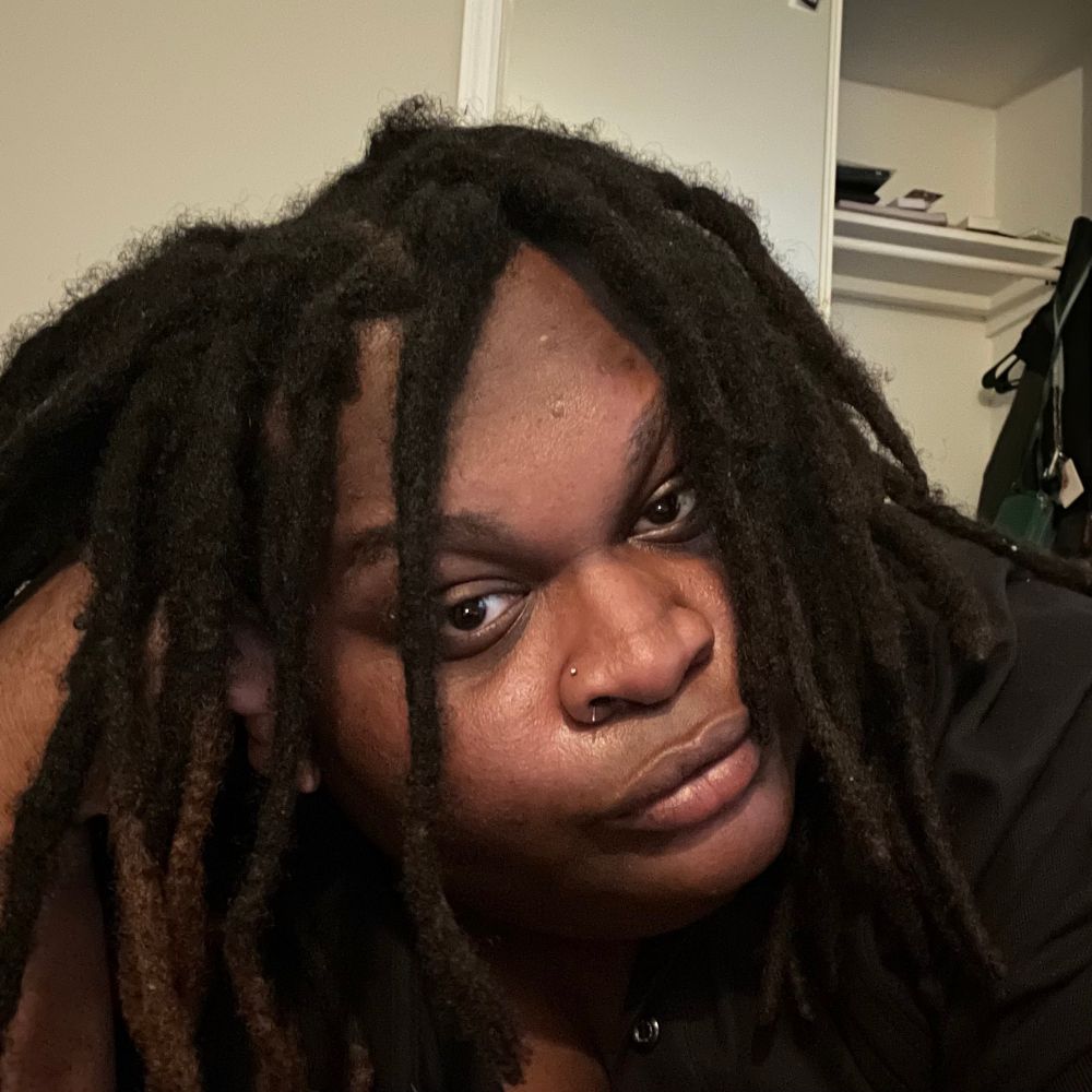 a selfie of myself. I am Black, brown skinned with thick shoulder length black locs. I have large facial features. I have high cheekbones, my cheeks are chubby, my lips and nose are big. I have a nose stud piercing on the left nostril of my nose. my eye color is dark brown. I have thick eyebrows. I am making a blank unassuming facial expression.
