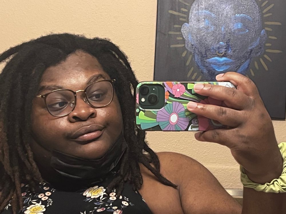 a selfie of myself. i have shoulder length semi freeform locs parted to the left, brown skin and black sleeveless dress that has white flowers on it. i have brown square framed glasses. i have a slight smile. my black mask is underneath my chin. i have a green scrunchie on my wrist and my phone has a colorful flowery case on it.