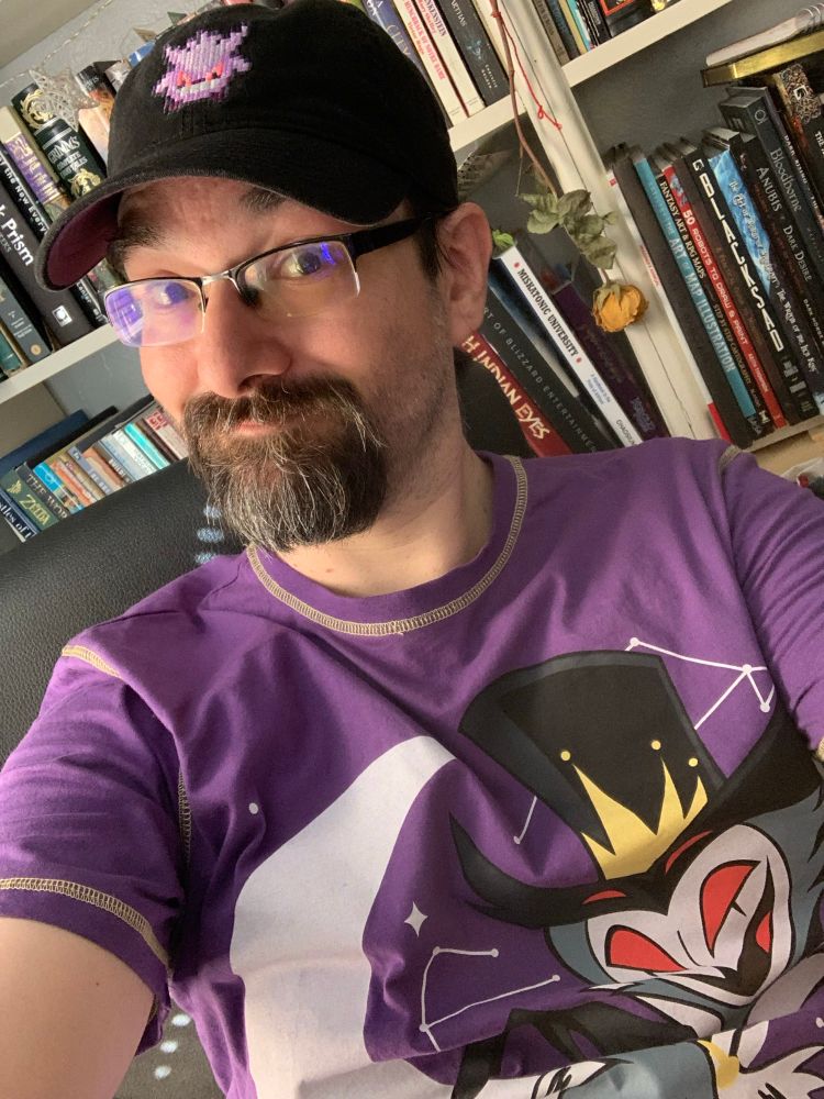 A dork (me) wearing a Stolas shirt from Helluva Boss & a Gengar Hat from Pokémon. Both purple and black/grey tones.