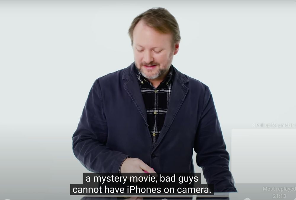 A still from a video showing Rian Johnson with light brown hair and a beard wearing a dark navy blazer over a black and white checkered shirt standing against a white background. He is speaking while looking down with a slight smile. The image has subtitle text at the bottom that reads "a mystery movie, bad guys cannot have iPhones on camera." 