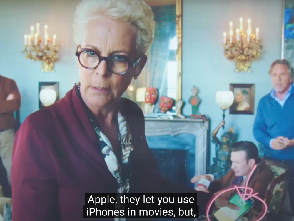 A still of a scene from the movie Knives Out showing Jamie Lee Curtis with silver blonde blonde hair and glasses, wearing a burgundy cardigan, standing in an elegant room with ornate wall sconces and a fireplace. She is speaking while holding an iPhone. Someone has circled the iPhone on the still in pink and drawn an arrow pointing at it. The image has subtitle text at the bottom that reads "Apple, they let you use iPhones in movies, but,"