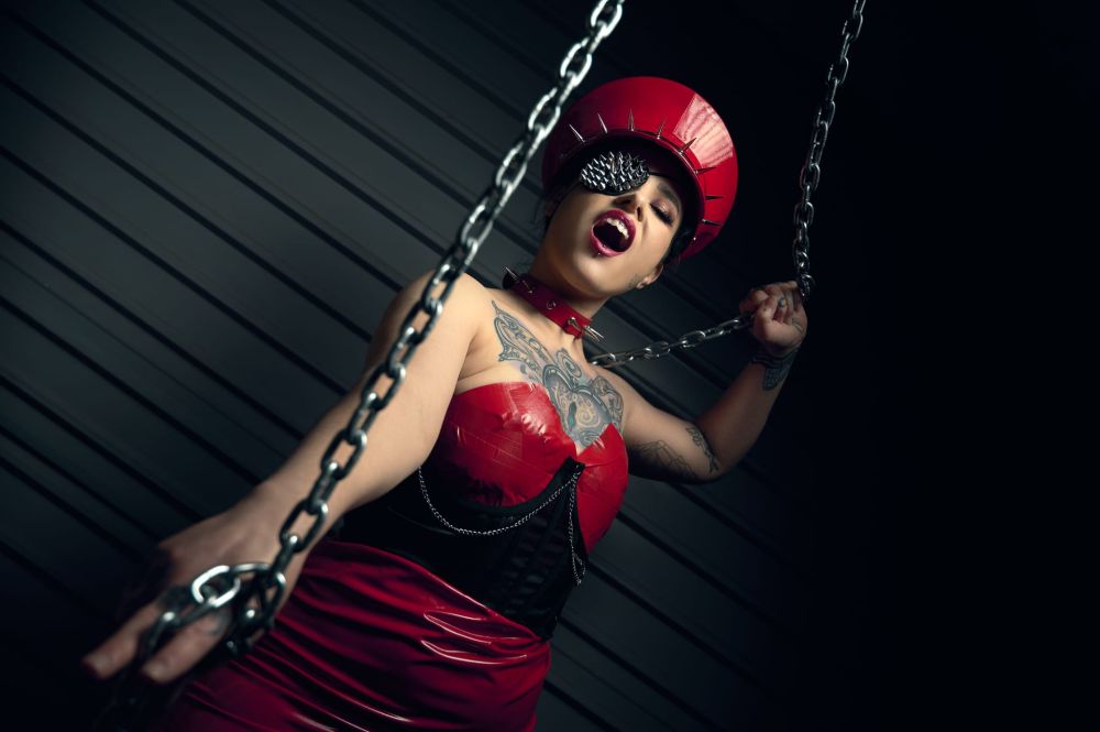 A tattooed Dominatrix looking into the camera with an imposing expression on their face stands in a dark room, wearing a Red PVC outfit holds onto chains suspended from the ceiling, motioning downwards with one hand, as to command one to get on the floor. 
#domme #prodomme #Dominatrix #findom #findomme #financialdomination #bdsm #skyhub #spicysky #swer #queer #queerswer #femelite #findomworld #findommeworld #🧡🖤 #Domination #Discipline #fetish #fetishist #kink #kinky #kinkster #fetlife #pvc #latex #militarycap #chains 