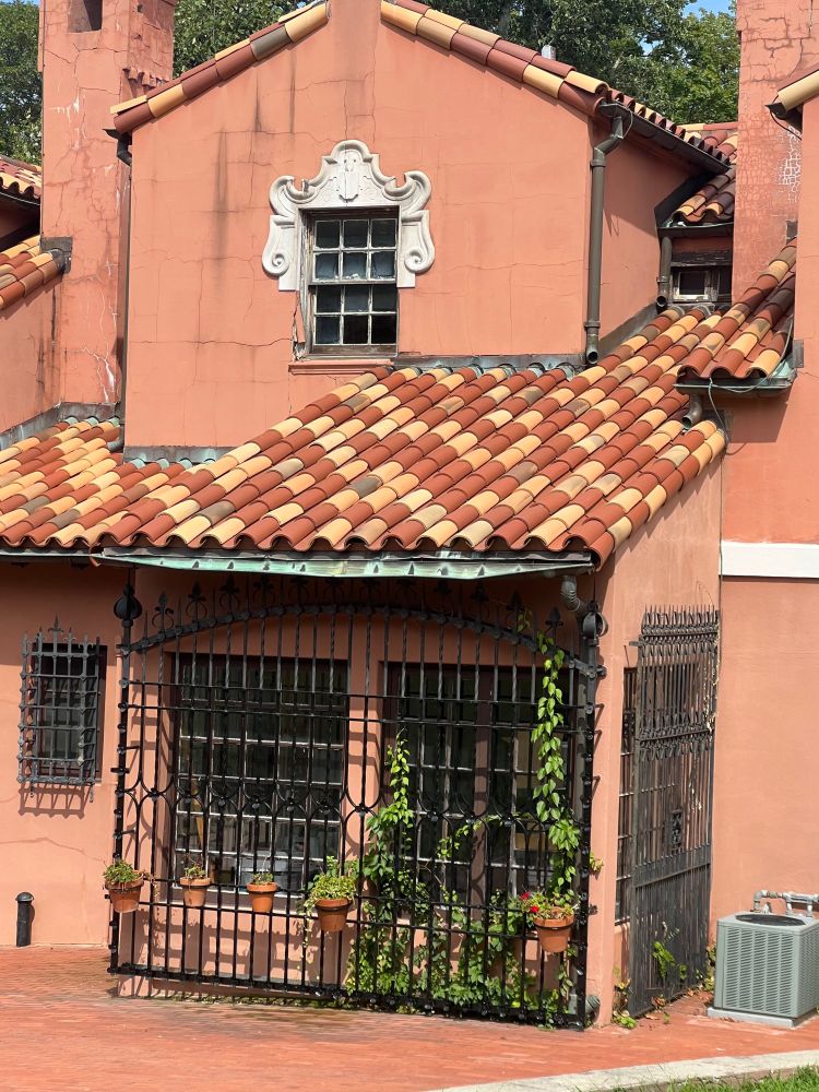 Picture of an orangey-pale orange(?) house, potted plants hung on a black fencing around the first floor, followed by very nice roof-tiles that speckle in colors from; reddish-brown, pale yellow, and a granite type. 