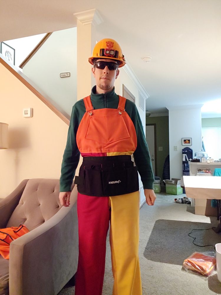 Man in a living room wearing a hard hat, safety glasses a green shirt and multicolor overalls (one leg red, one leg yellow, and an orange bib)