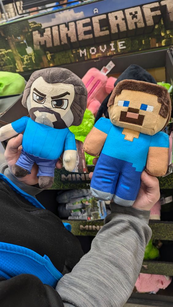 In this picture we are at a minecraft movie feature at a walmart. my partner is holding two steve plushies. One being from the game itself and the other from the movie. The movie plush is literally just jack black with a blue shirt on lol