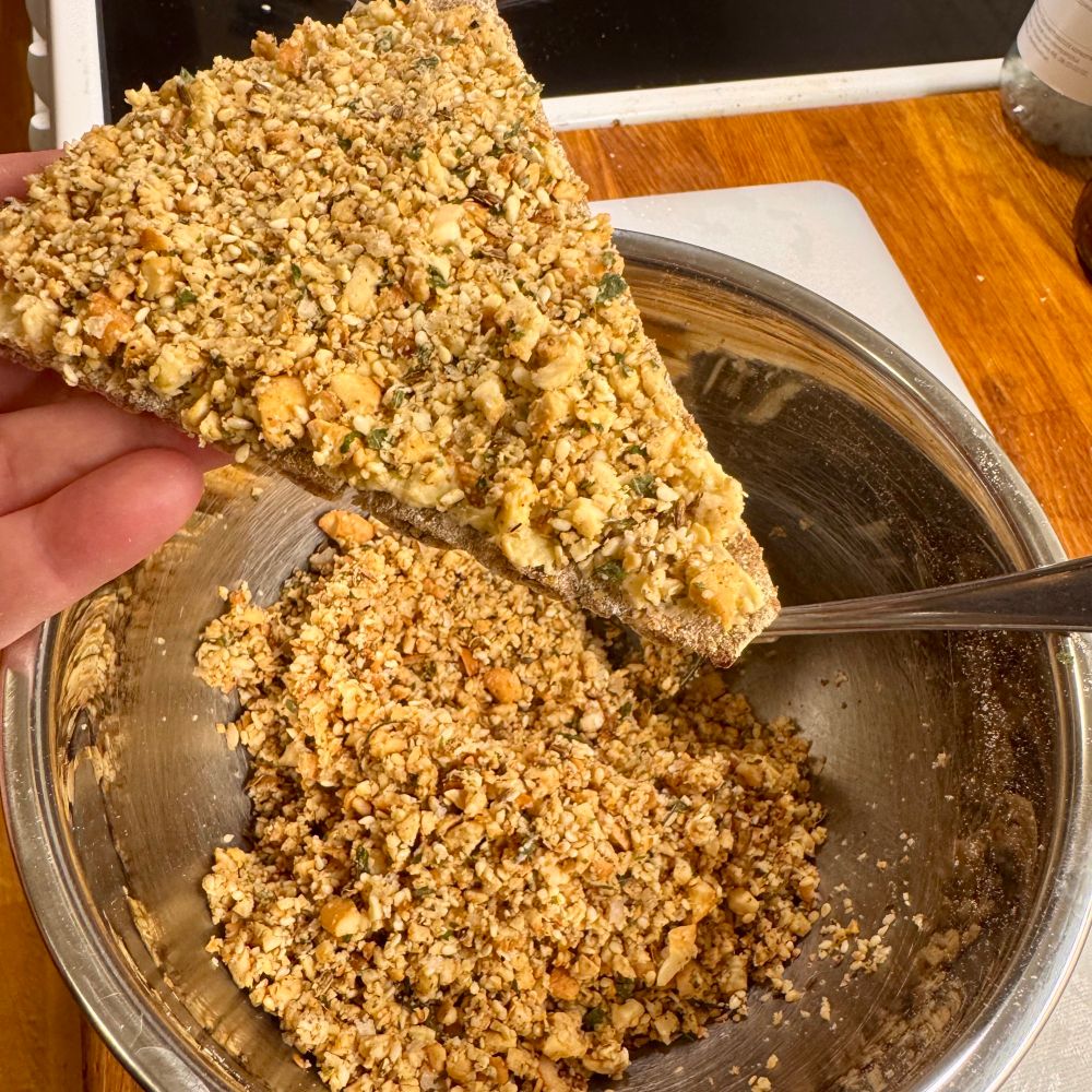 På en köksbänk står en metallskål som innehåller nöt, frö och kryddblandningen. Ovanför skålen håller jag ett trekantigt knäckebröd med hummus och blandningen på. 