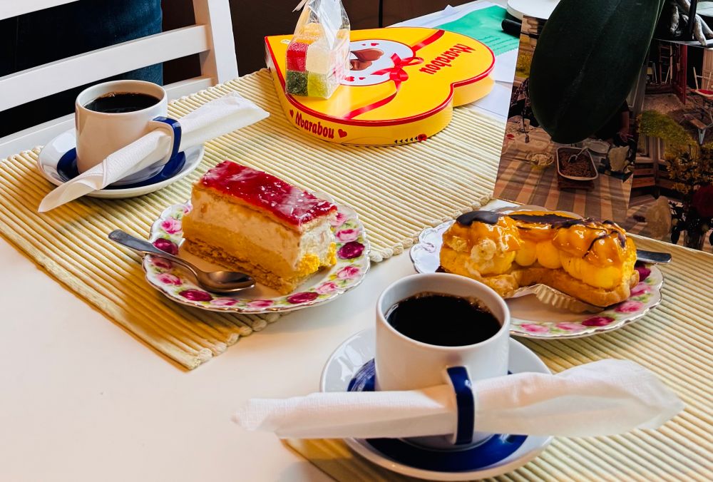 Detaljbild på köksbord: starkt, svart kaffe i två rejäla kaffekoppar, två bakelser på varsin assietter med rosenmönster. I bakgrunden skymtar en påse med marmeladgodis och en marabouask med chokladhjärtan. 
