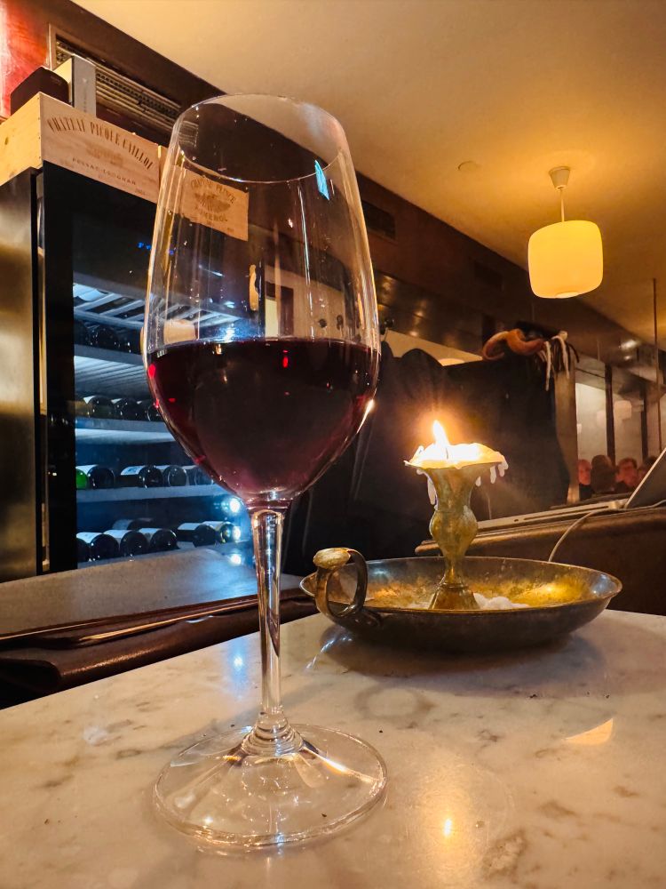 Ett halvdrucket glas rödvin på en bardisk, ett nedbrunnet ljus i en stor mässingsstake står snett bakom. En full vinkyl skymtar i bakgrunden. 