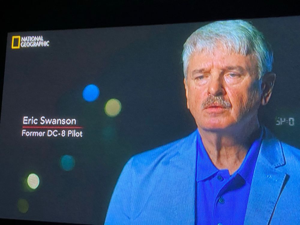 ひとつ前の写真と同じく、25/06/14の0時よりナショジオで放映された「メーデー！」(s22/エア・トランスポート・インターナショナル782便)のテレビ画面を撮影した写真。青いシャツ・水色のジャケットを着て、短いグレイヘアーの白人男性が喋っているところ。その横に字幕で「Eric Swanson」、改行して「Former DC-8 pilot」と表示されている。