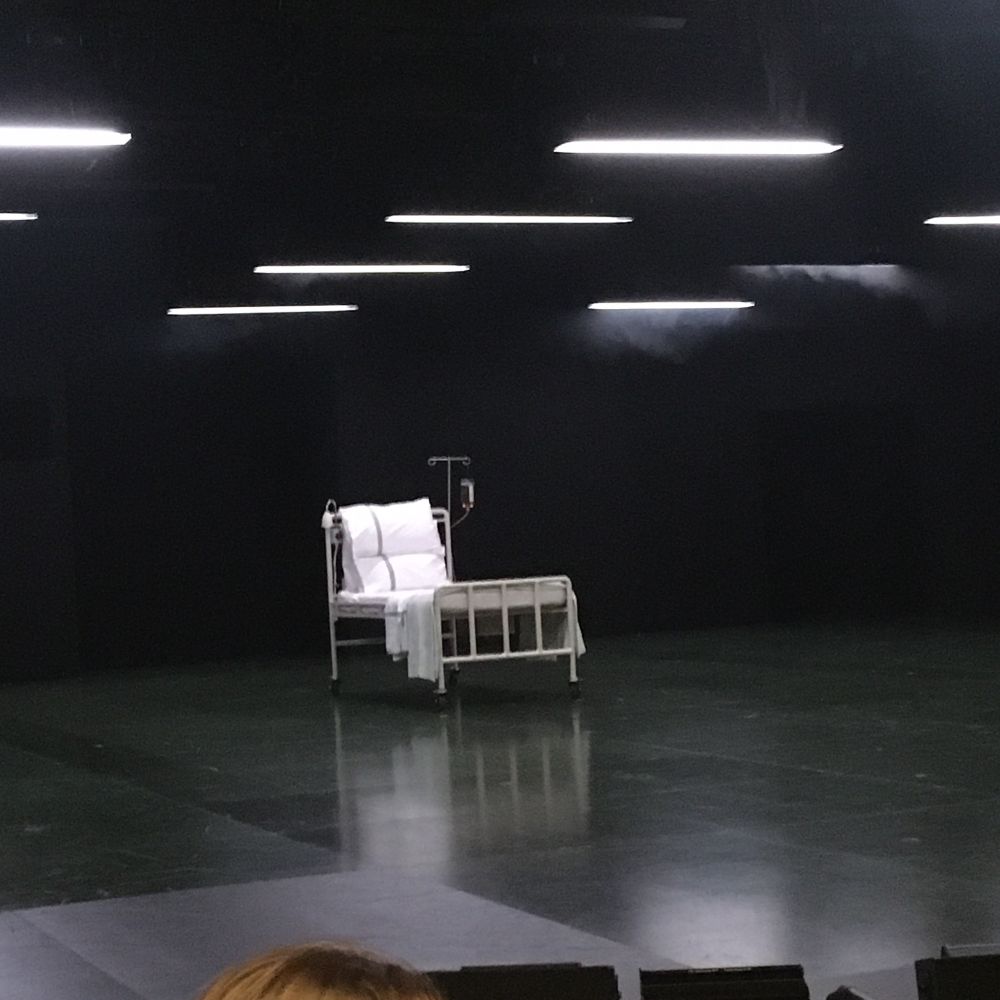 Empty dark stage except for a 1950s NHS bed