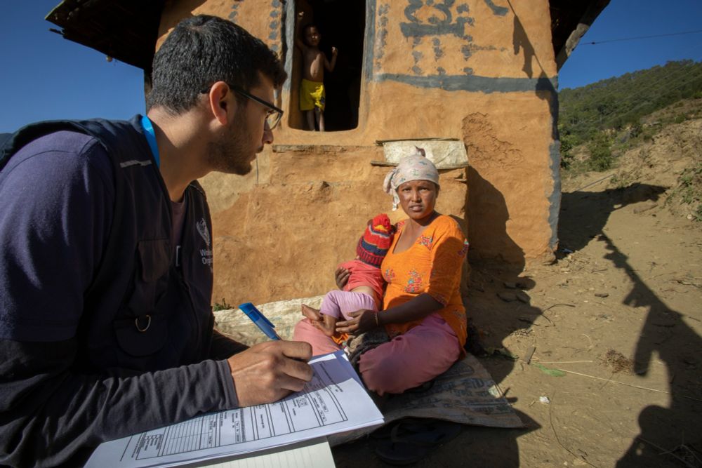 Picture of a WHO staff member conducting a survey in a rural area. He is holding pen and paper and talking to a woman who sites outside her hut. She has a toddler in her arm, another child stands in the background.