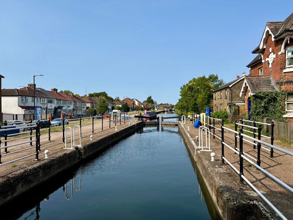 Photo showing a canal lock in an urban area. There are railings on both sides of the lock, and low rise houses beyond, with trees dotted about, and some cars parked beside the road to our left. The sky is pale blue, and a darker version of it is reflected in the calm canal water.