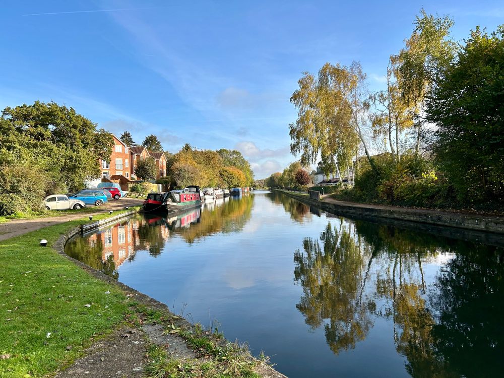 Photo of a canal, taken on a sunny autumn day. The bank of the canal bends away slightly on the left, and is mostly grassy. Continuing up the left bank, we see some parked cars and some houses, and narrowboats (barges) moored beside; further on we see trees with autumn leaves, and all of this is lit by bright sunshine. Meanwhile, the right bank has tall trees and bushes, and is mainly in shadow. The water of the canal is still and acts like a mirror, giving an almost perfect reflection of the trees, sky, narrowboats and houses above.  The sky is blue, with some high vaporous clouds, plus some puffy white clouds in the far distance.