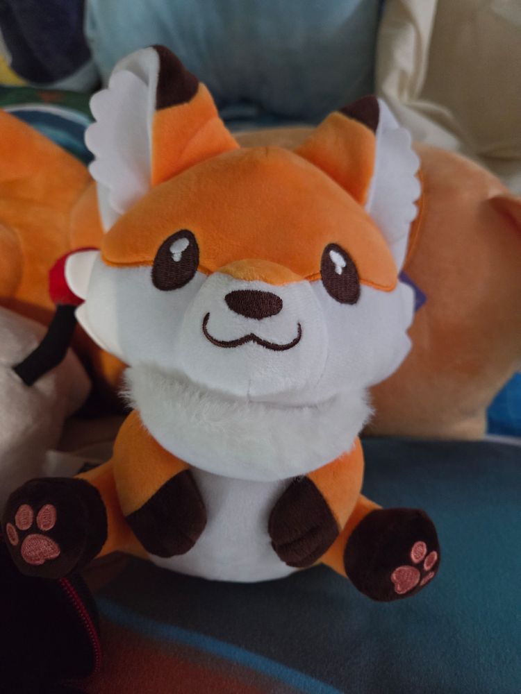 Vicksy Sitting Plush comfortably on a big pillow and surrounded by plush friends