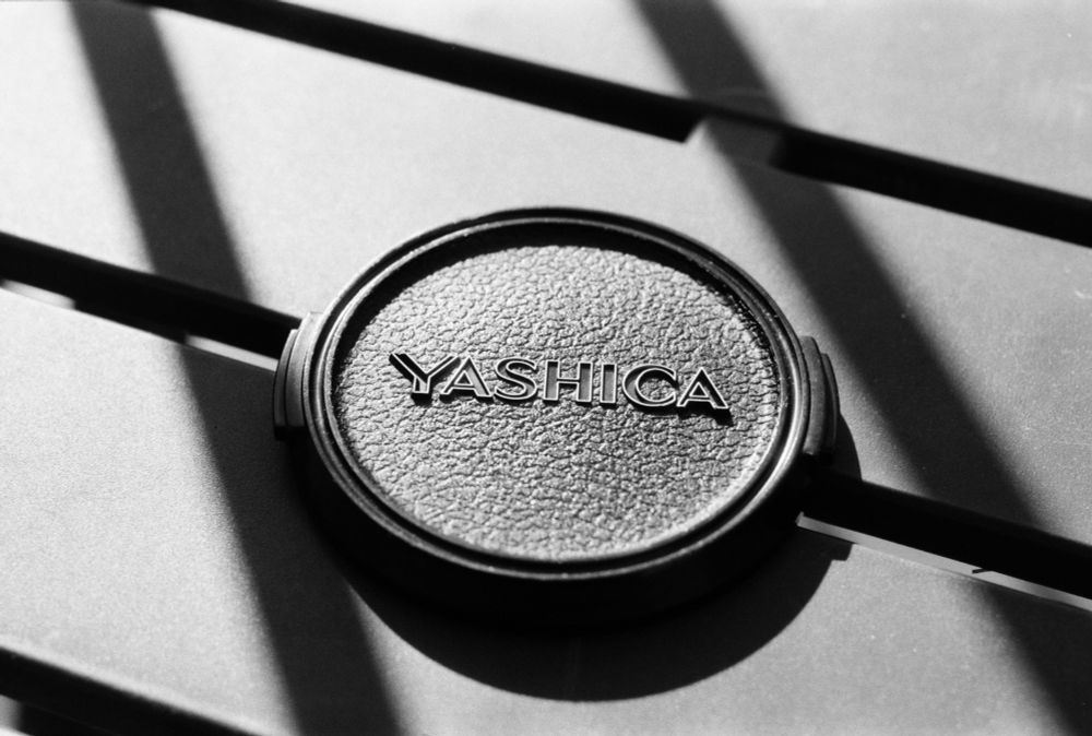 black & white: close up of a YASHICA lens cap on a table with two parallel diagonal shadows either side of it