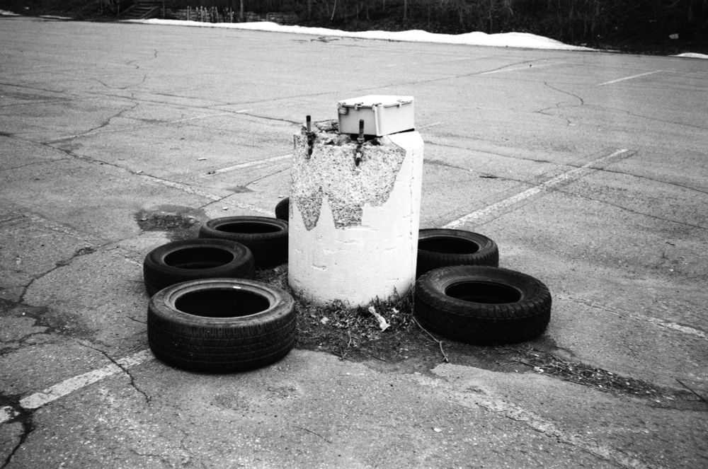 black & white: a ring of tires surrounding the base of what would be a street lamp in the middle of a parking lot but it’s just the base and there’s like an electrical box on top of it and one of the tires is missing in the foreground so the ring isn’t complete