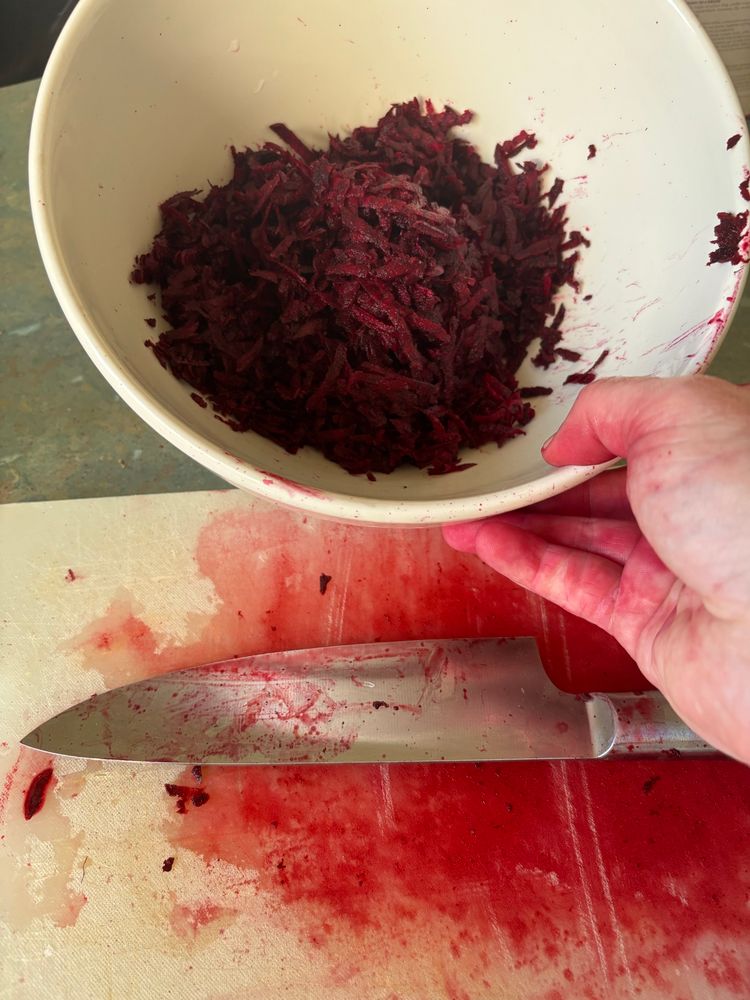 A bowl of grated beetroot and a beet stains knife and chopping board and my hand and is that some the ceiling
