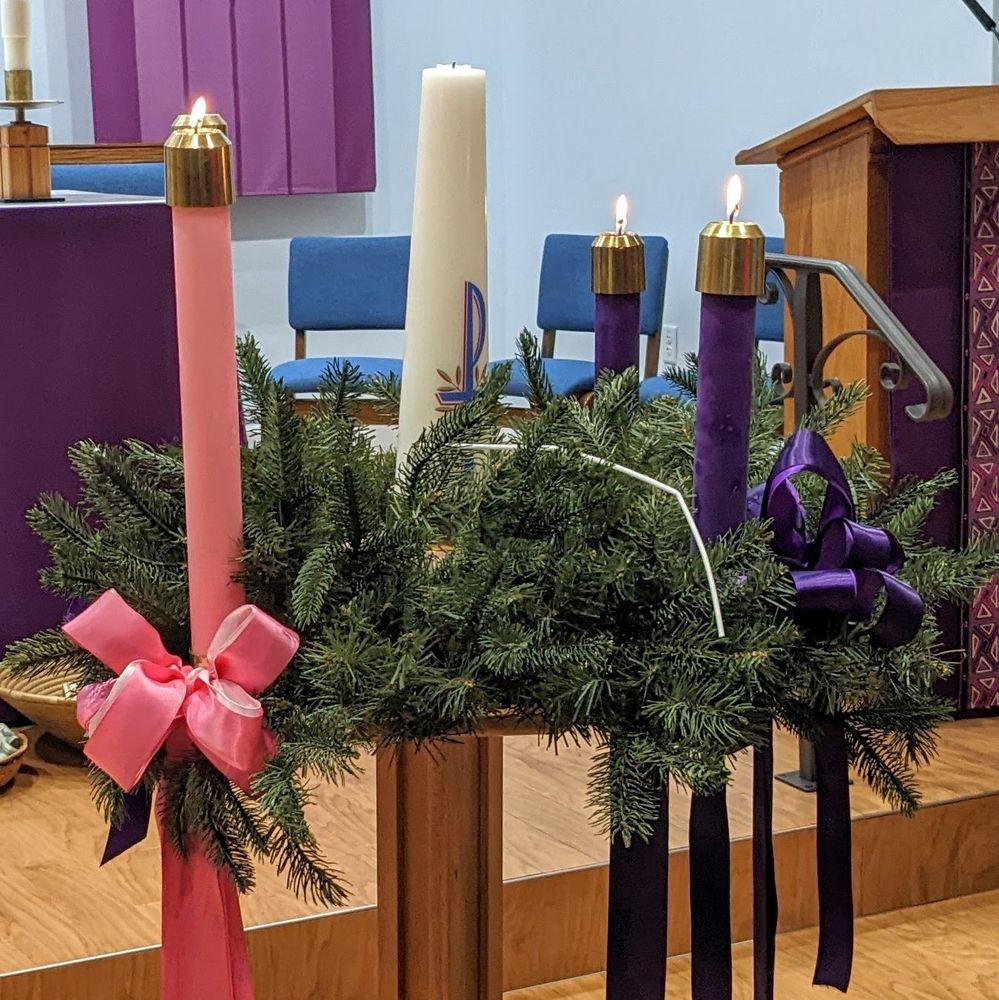 an advent wreath with 3 candles lit, 2 purple and one pink, each seated among evergreen garland surrounding a white pascal candle