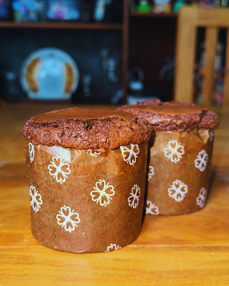 foto de dois brownietones (brownie no formato de panetone) lado a lado sobre uma mesa de madeira. ao fundo e desfocado, uma estante com decorações de casa