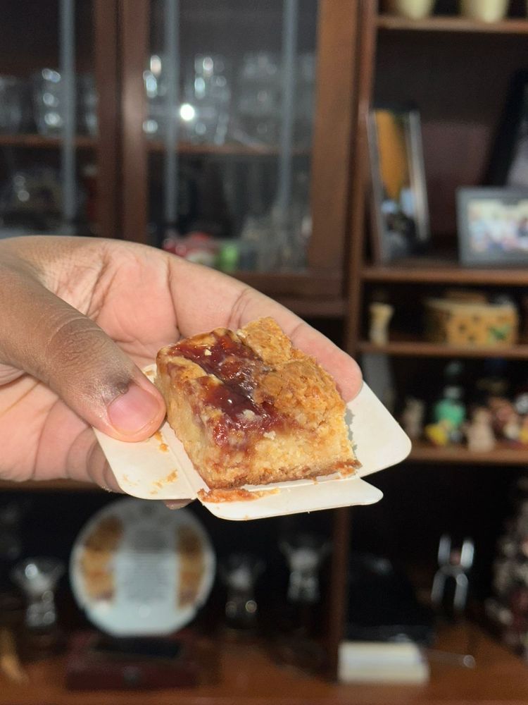 foto de uma mão negra segurando uma fatia de brownie romeu e julieta (de queijo e goiabada). a fatia é de massa clara, levemente bronzeada, com uma generosa camada de goiabada derretida por cima e no meio da massa.