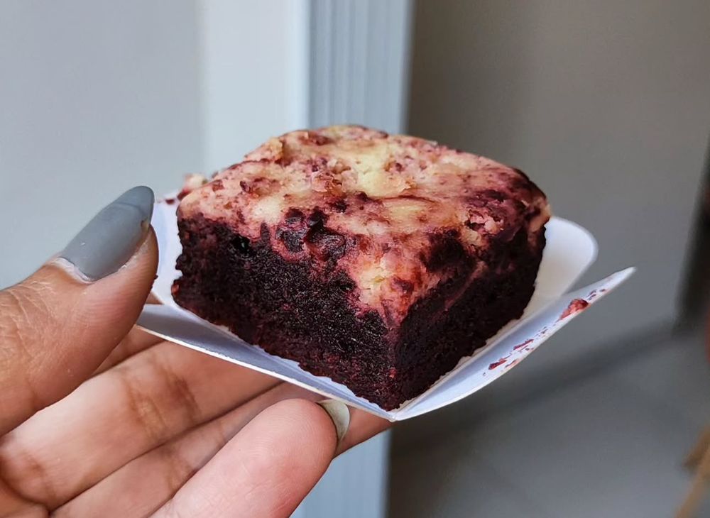 foto de uma mão segurando uma embalagem branca que contém um brownie sabor red velvet. a massa do brownie é vermelho escura e, em cima, uma camada mesclada creme nas cores branco e vermelho)