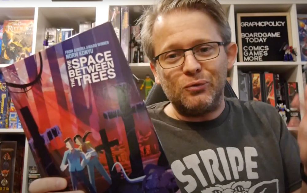 Brett holds a copy of the Space Between the Trees to the camera backed by a truly impressive comic collection