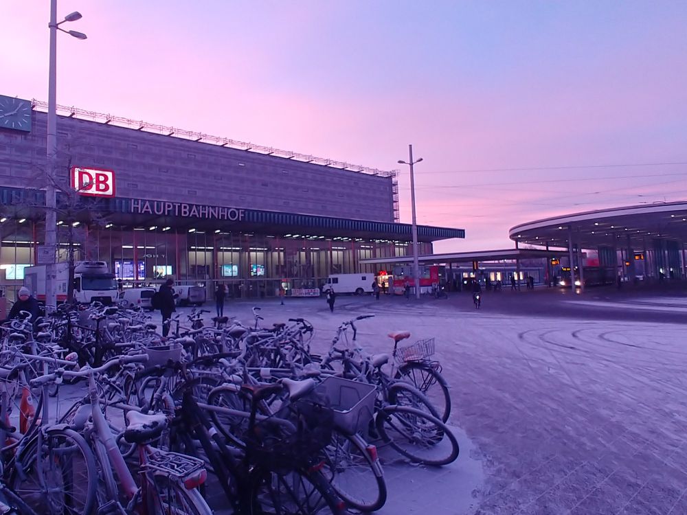 A picture of Braunschweig rail station building and bike parking covered with snow and bathed in a warm orange and lilac sunrise light.