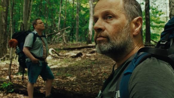 screen shot from "good one" (india donaldson, 2024). two male characters are in the woods. one is in the foreground, another is in the background. they are wearing backpacks. they are terrible, ordinary men.