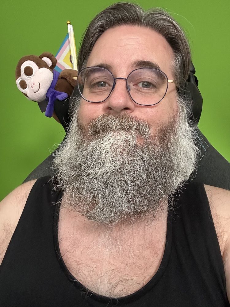 Me wearing a black tank top and glasses with blue circular frames. Over my right shoulder, brown plush Chair Monkey has been joined by the intersex-inclusive pride flag (a flag with horizontal stripes in red, orange, yellow, green, blue and purple, with an arrow made up of black, brown, light blue, pink and white stripes with the remaining triangle filled with yellow containing a purple circle)