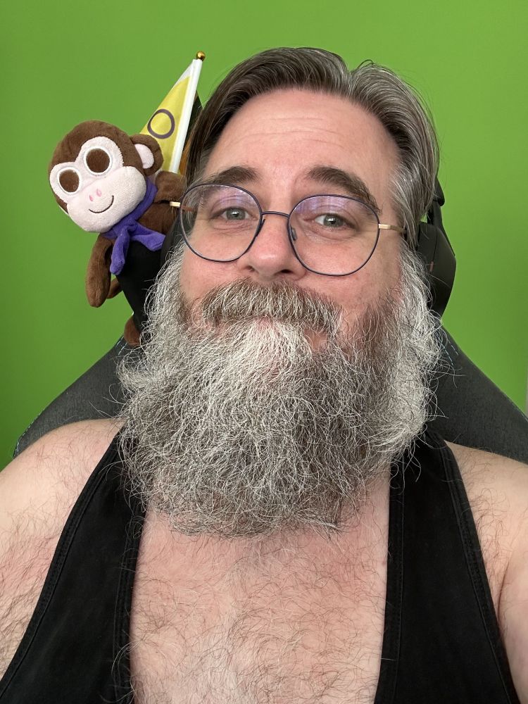 Me, a scruffy grey-bearded man wearing a black tank top and blue-rimmed glasses, seated in front of a green screen with brown plush Chair Monkey and a progress pride flag attached to the chair above my shoulder