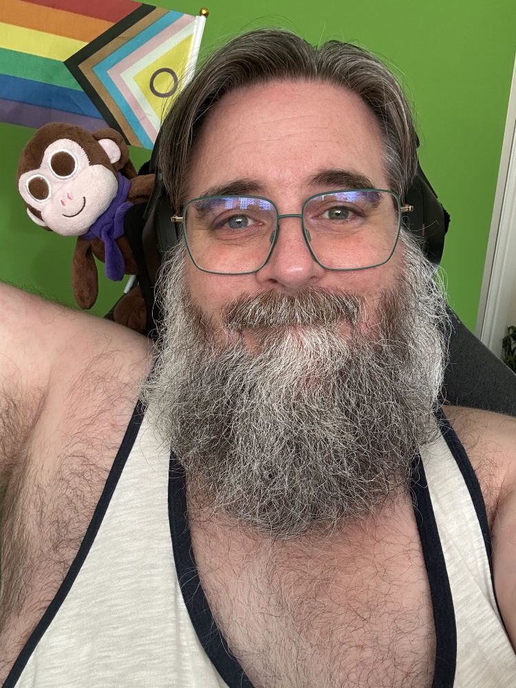 Me wearing a cream tank top and square green-rimmed glasses with brown plush Chair Monkey and the progress pride flag over one shoulder on the back of the chair