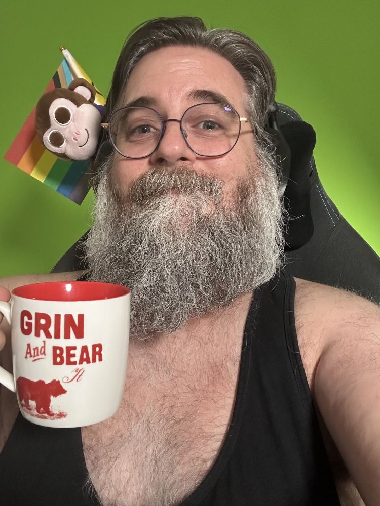 Me wearing a black tank top and holding a white mug with the text "Grin and Bear it" and the silhouette of a bear in red. Brown plush Chair Monkey peeps from the back of the chair behind me with the progress pride flag