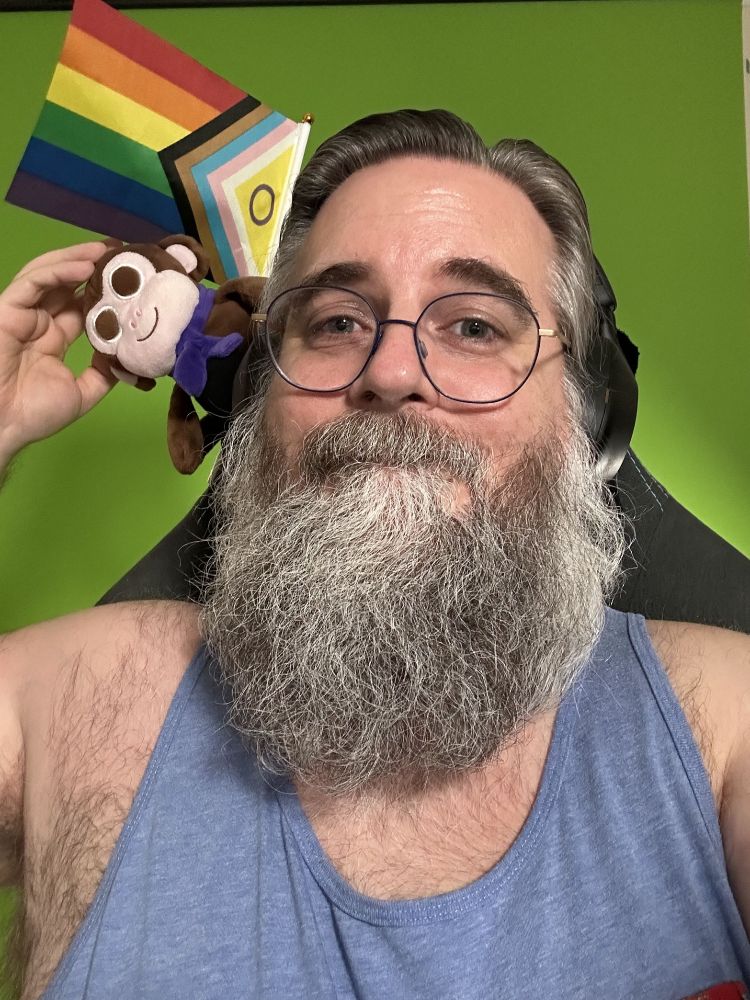 Me wearing a blue tank top, reaching to reposition brown plush Chair Monkey flopping sideways from the back of my chair just below a pride flag