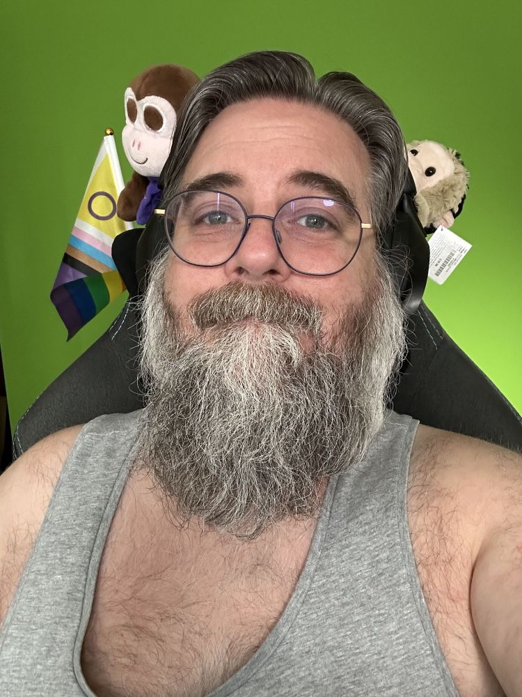 Me wearing a grey tank top. Over my left shoulder, black and white plush Chair Monkey is partially visible, flopping from the back of the chair. Over my right shoulder, brown plush Chair Monkey has been joined by the intersex-inclusive pride flag (a flag with horizontal stripes in red, orange, yellow, green, blue and purple, with an arrow made up of black, brown, light blue, pink and white stripes with the remaining triangle filled with yellow containing a purple circle)