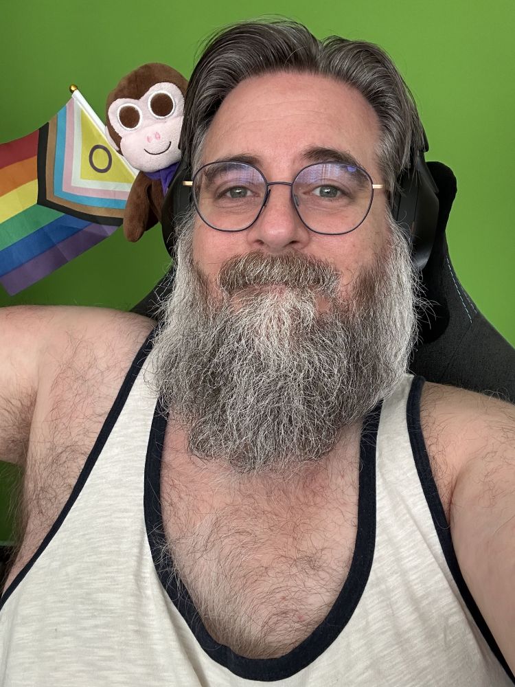 Me wearing a cream tank top and glasses with blue circular frames. Over my right shoulder, brown plush Chair Monkey has been joined by the intersex-inclusive pride flag (a flag with horizontal stripes in red, orange, yellow, green, blue and purple, with an arrow made up of black, brown, light blue, pink and white stripes with the remaining triangle filled with yellow containing a purple circle)