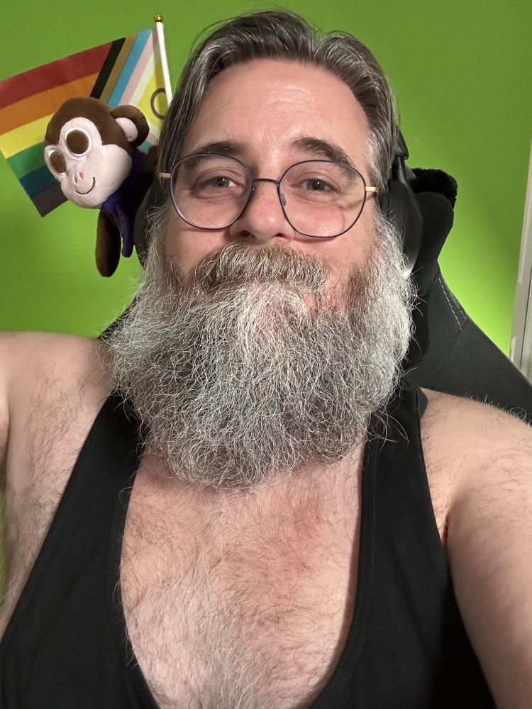 Me wearing a black tank top and glasses with blue circular frames... and looking warm. Over my right shoulder, brown plush Chair Monkey has been joined by the intersex-inclusive pride flag (a flag with horizontal stripes in red, orange, yellow, green, blue and purple, with an arrow made up of black, brown, light blue, pink and white stripes with the remaining triangle filled with yellow containing a purple circle)