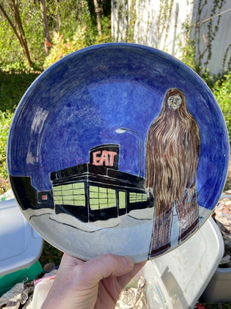 A ceramic plate with an underglaze painting of a snowy scene with Bigfoot standing outside a diner at night