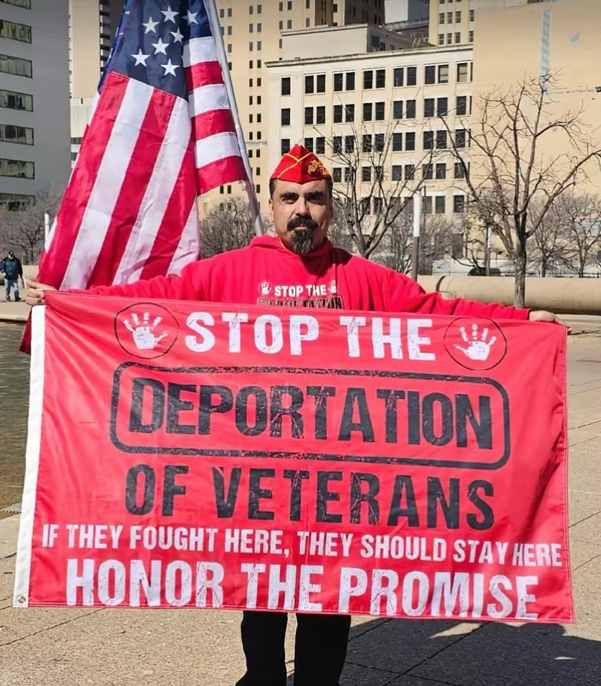 Man holding a U.S. flag and a red banner that says, “Stop the deportation of veterans. If they fought here, they should stay here. Honor the promise.”