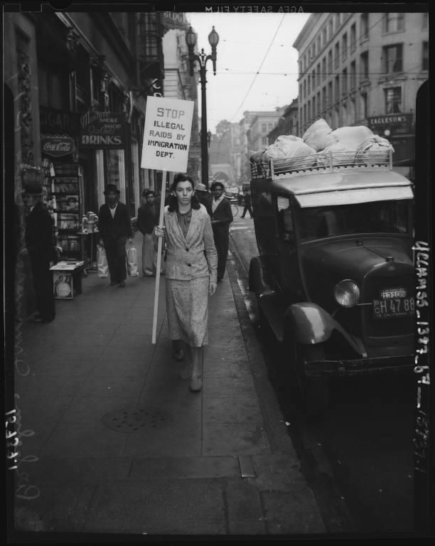 Black-and-white photo of a woman protesting on a city sidewalk in 1930s Los Angeles. She holds a sign that reads, “STOP ILLEGAL RAIDS BY IMMIGRATION DEPT.” A vintage car loaded with sacks is parked nearby, and several men stand in front of a store watching her pass.