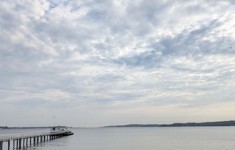 Meer mit Bootssteg, stark bewölkter Himmel, leichter Schimmer von Morgensonne am rechten Bildrand.