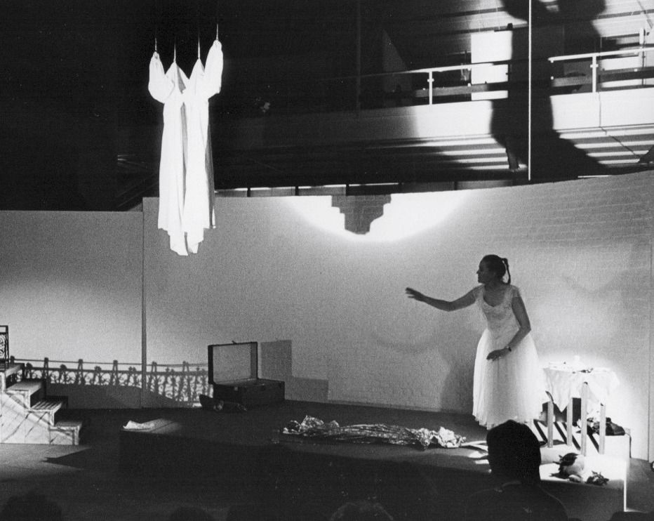 Eine Bühne mit abstrakten Elementen und einer weißen, lang gekleideten Darstellerin, die die Hände ausstreckt, während das Publikum im Hintergrund sitzt.