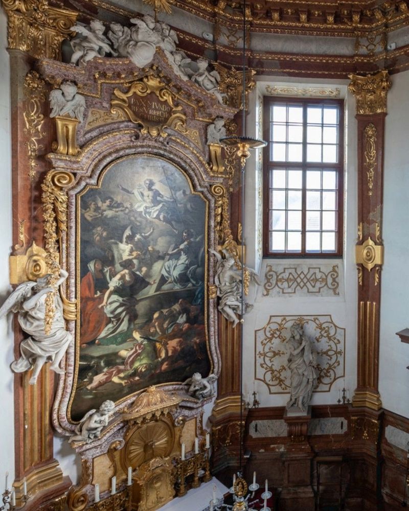 An ornate and lavishly decorated interior with a large, gilded altarpiece featuring a religious painting amid statues, carvings, and architectural details.