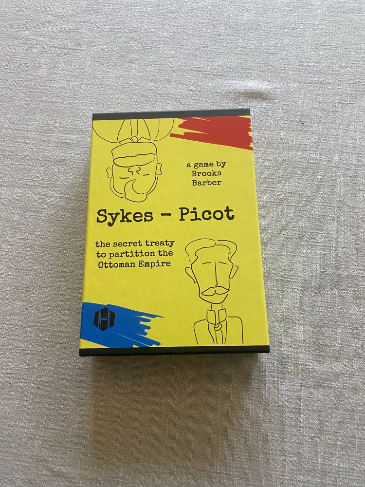 A copy of the board game, Sykes-Picot, designed by Brooks Barber and published by Hollandspiele. A black box wrapped in a yellow cover. In the corners of the cover are red and blue marks resembling marker strokes. In the other corners are two doodles of the eponymous Sykes and Picot, drawn by Amabel Holland. The title is in the center of the box, with the designer’s name to the right and to the left the subtitle “the secret treaty to partition the Ottoman Empire.”