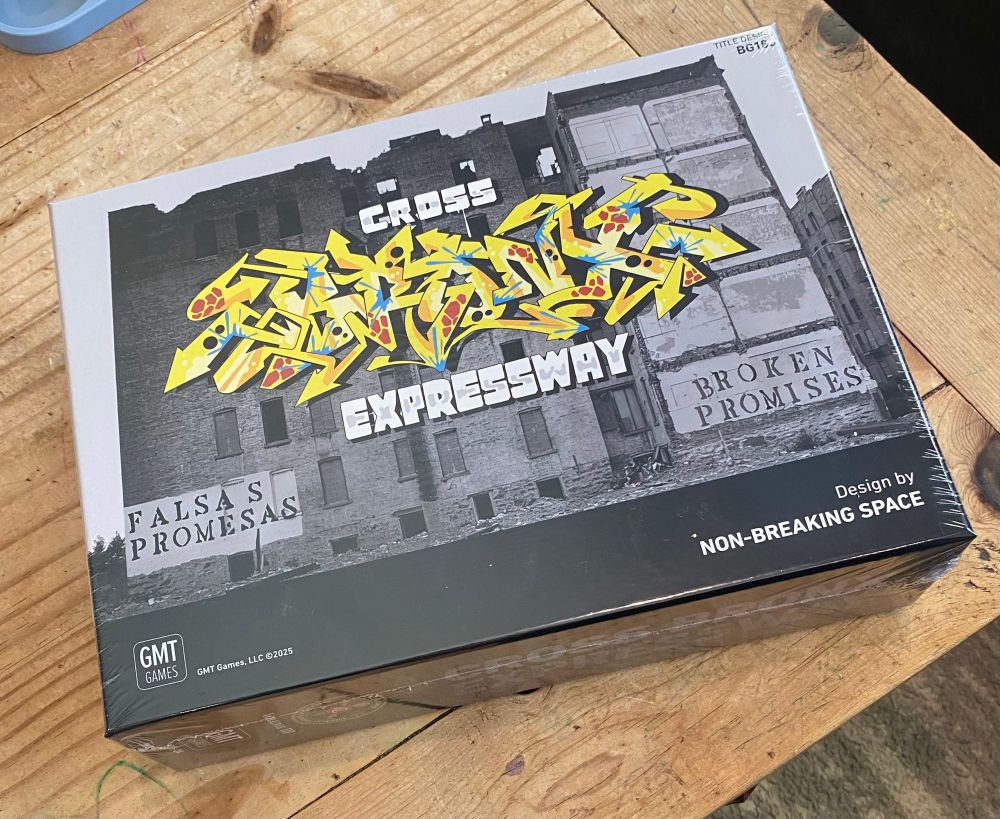 A board game laying on a wooden coffee table, slightly askew. The cover shows a dilapidated build on in the South Bronx. In the bottom left corner is written “FALSAS PROMESAS” and in the bottom right “BROKEN PROMISES.” Across the middle of the cover is the title Cross Bronx Expressway. Cross and Expressway are in reserved black and white lettering and the word Bronx is in yellow, blue, and red graffiti style text. 
