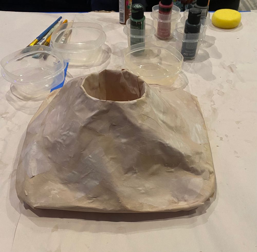 A table covered in paper with a papier-mâché volcano resting in the middle. Some paints, brushes, and containers in the background. 
