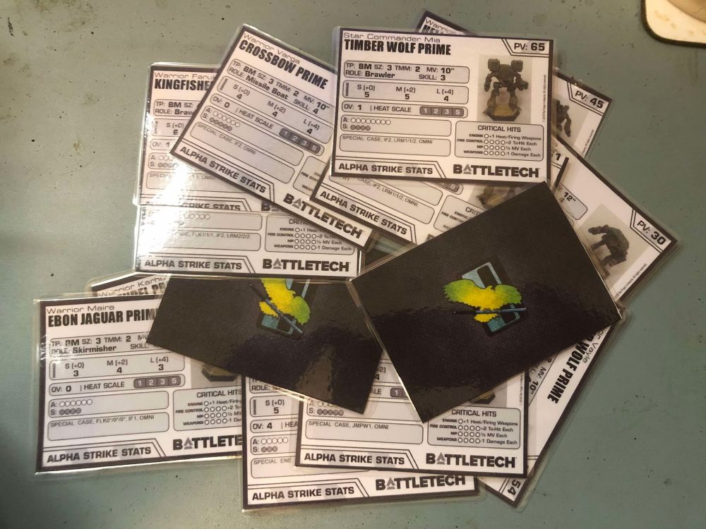 A photo of a pile of Alpha Strike stat cards for Clan Jade Falcon 'mechs. They are card which has been laminated. One side has a photo of a miniature with the relevant stats for the game, the other is black with the Clan Jade Falcon emblem in the centre.