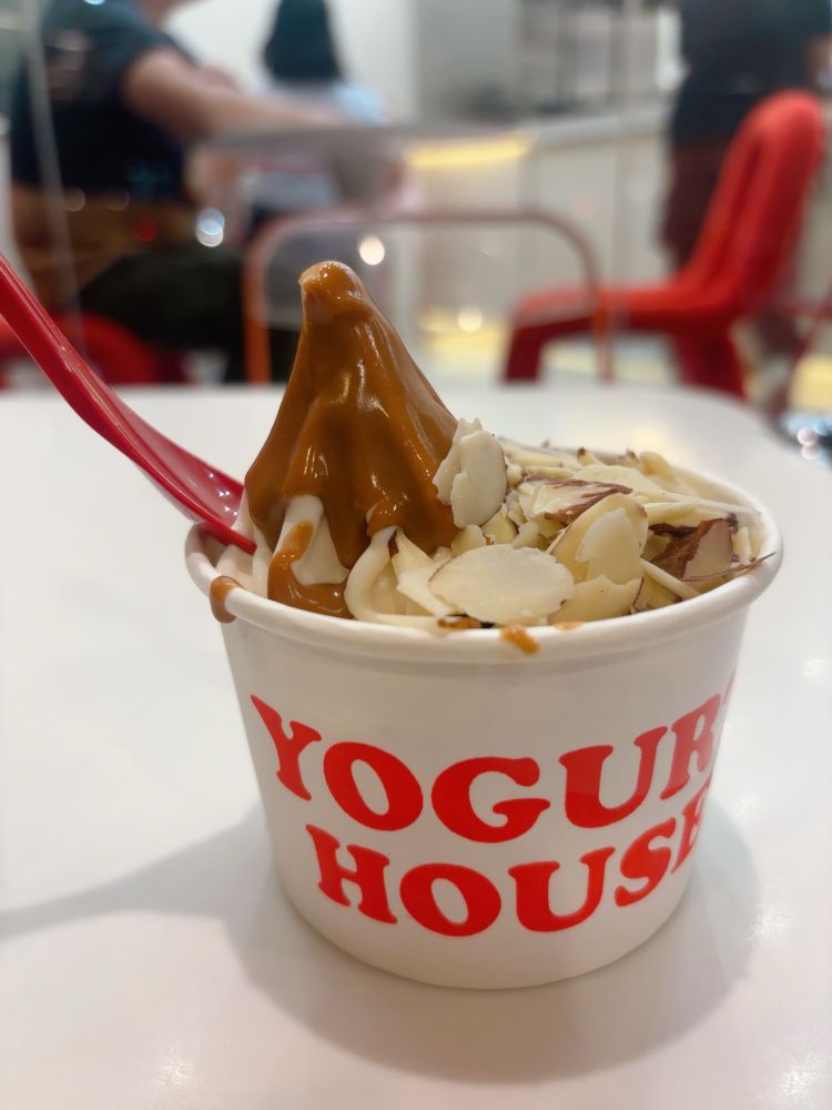 白いカップに盛られたフローズンヨーグルトの上にソースとスライスアーモンドがトッピングされている。赤いスプーンが添えられており、背景には椅子や人影がぼんやり映っている。