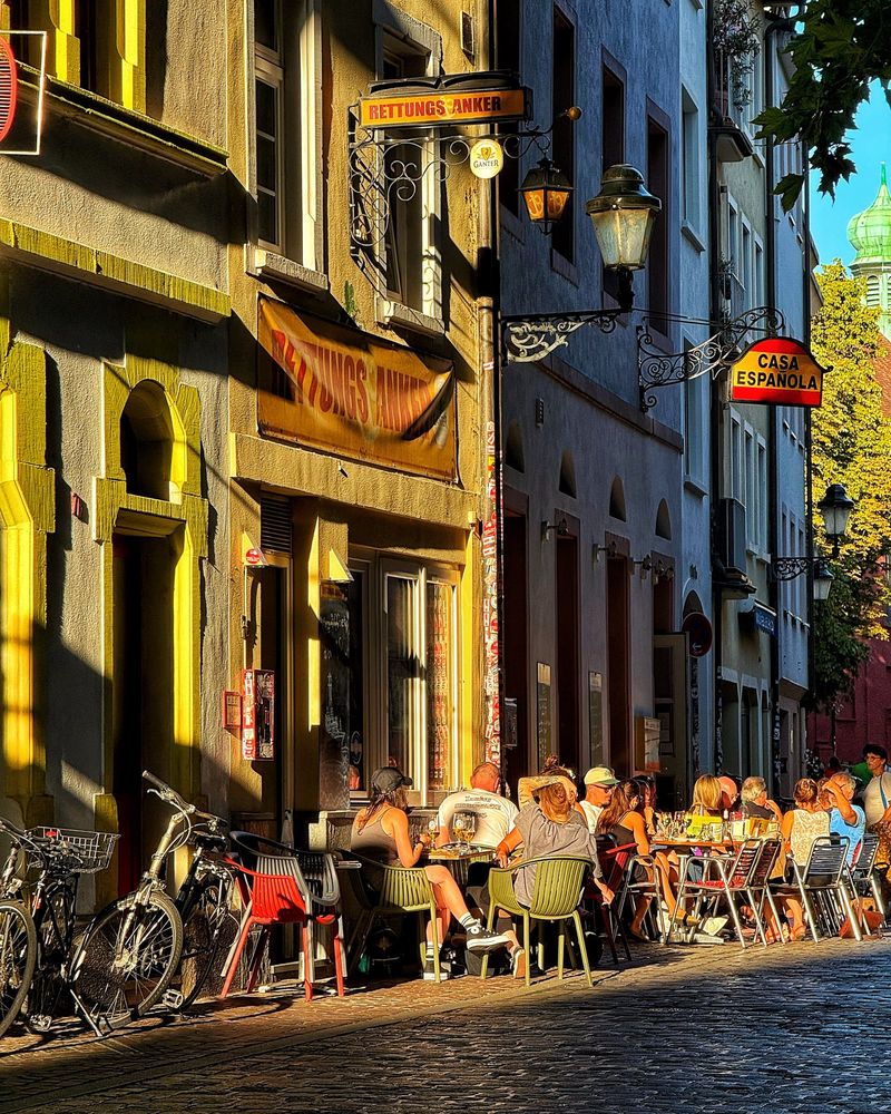 A street scene in an old part of town. Low sunlight beams through the street, casting shadows and warm tones, accentuating the randomly angled building facades. In the main strip of sunlight, people sit outside cafes and restaurants, some of whose names are hung on signs.