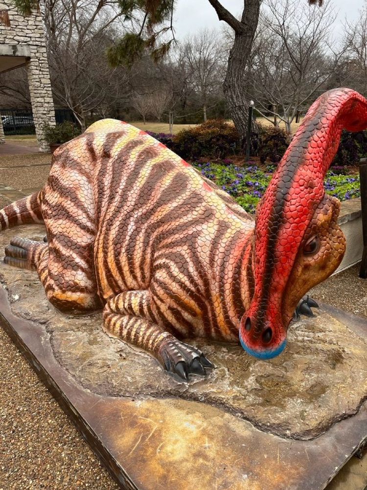 One of the dinosaur exhibits featuring a Parasaurolophus with a red head, blue lips, and tiger stripes on body. It is prostrate with chest to ground, as if welcoming guests to jump on its back!