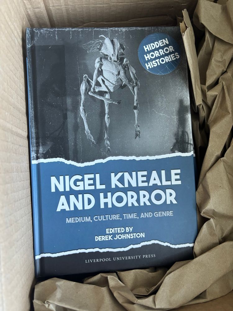 An open cardboard mailing box shows, nestled in brown packing paper, the cover of the book “Nigel Kneale and Horror”. The cover is in blue and grey tones, a broad band displaying the title, a circle showing the Hidden Horror Histories series title, and a greyscale image showing a hovering, three-legged, three-armed insectile Martian from “Quatermass and the Pit”.
